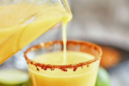 A yellow drink being poured into a cocktail glass with a peppered rim